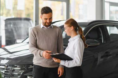 Araba satıcısı, otomobil galerisinde gülümseyen bir erkek müşteriye bir belge sunuyor.