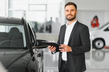 Modern galeride gülümseyen takım elbiseli genç profesyonel araba satıcısı satış yardımı ve müşteri hizmetleri uzmanlığı teklif ediyor.