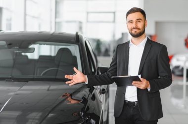 Resmi kıyafetli profesyonel satıcı modern bir galeride müşterilere yardım eden siyah bir araba sunuyor.