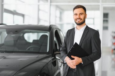 Genç adam gülümsüyor, pano tutuyor, modern bir galeride arabanın yanında duruyor, mükemmel bir hizmet sunuyor.