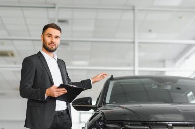 Satıcı elinde bir pano, modern bir galeride siyah bir araba sunuyor, otomobil alımı hakkında tavsiyelerde bulunuyor.