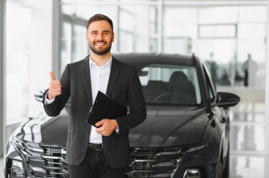 Profesyonel bir adam gülümsüyor, elinde bir not panosu var ve yeni bir otomobilin yanında baş parmağını kaldırıyor.