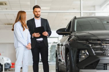 Takım elbiseli satıcı, kadın müşteriyle, modern, parlak otomobil galerisinde, satın alma kararlarında danışmanlık yaparak, pano üzerinden araba özelliklerini tartışıyor.