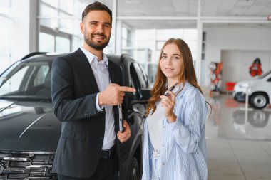 Profesyonel araba satıcısı yeni bir aracı işaret ederek modern bir galeride gülümseyen bir kadın müşteriye arabanın anahtarlarını veriyor.