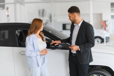 Satıcı, bir araba galerisinde kadın bir müşteriyle araç özelliklerini ve şartları tartışıyor ve satın alma sürecine odaklanıyor.