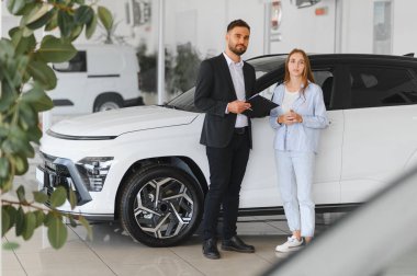 Modern bir araba galerisinde kadın müşteriye danışmanlık yapan satıcı, araba özelliklerini ve satın alma seçeneklerini tartışıyor.