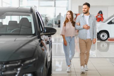 Genç mutlu çift araba galerisinde yürüyor, yeni araçları keşfediyor ve birlikte bir araba almayı planlıyor.