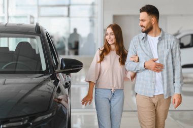 Genç çift kol kola yürüyorlar, bir otomobil bayisinde siyah bir arabaya bakıyorlar, satın alma kararı veriyorlar.