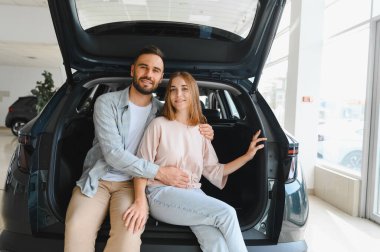 Mutlu genç çift, bir galerideki yeni beyaz bir cipin bagajında oturuyor. Gülümsüyor ve araba satın alma kararını kutluyorlar.