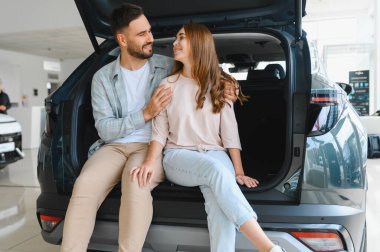 Modern bir cipin bagajında oturan genç, gülümseyen bir çift, bir araba galerisinde birbirlerine bakıyorlar.