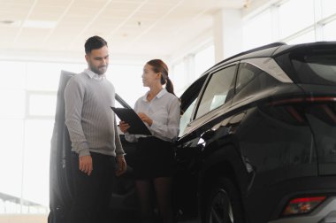 Satıcı ve erkek müşteri, modern bir galeride koyu renk bir SUV 'un alım veya kiralama seçeneklerini tartışıyor.