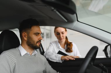 Arabada oturan bir erkek müşteri bir otomobil galerisinde kadın bir satıcıyla pazarlık ediyor ve satın alma kararı veriyor.