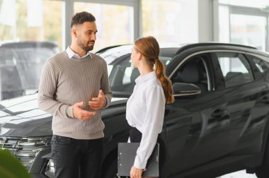 Kadın ve erkek araba galerisinde durup otomobil özellikleri ve seçenekler hakkında konuşuyorlar. Satış yardımcısı pano tutuyor