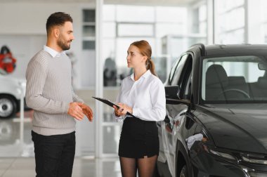 Satıcı kadın elinde bir pano ve bir erkek müşteriyle otomotiv galerisinden yeni bir araç almakla ilgili konuşuyor.