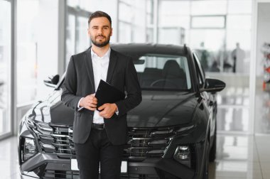 Satıcının elinde belgeler var, kameraya gülümsüyor, otomobil galerisinde modern siyah bir otomobilin önünde duruyor.