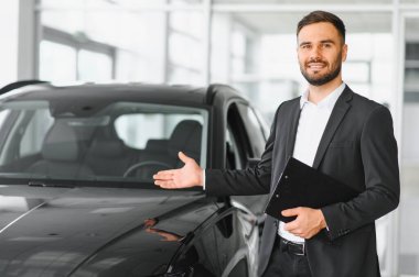 Takım elbiseli profesyonel satış elemanı, parlak, modern bir galeride parlak, siyah bir araba sunuyor.