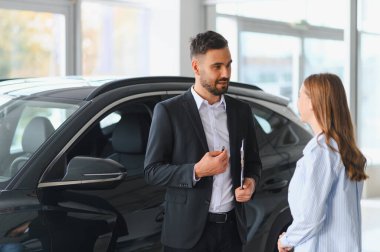 Yeni bir aracın yanında duran araba satıcısı modern bir galerideki kadın müşteriye detayları anlatıyor.