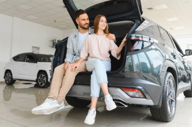 Genç mutlu çift yeni arabanın içini keşfediyor, modern galeride açık sandıkta oturuyor, gülümsüyor.