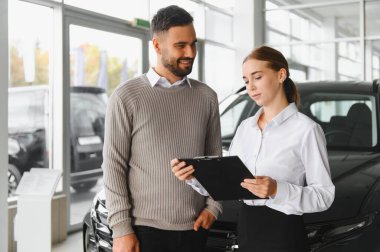 Satış elemanı, bir erkek müşteriye evrak işlerinde yardım ediyor, otomotiv perakendecisinde araba alımını tartışıyor.