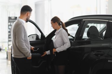 Satıcı kadın, müşteriye yeni bir otomobil seçmesinde yardımcı oluyor, modern bir araba galerisinde özellikleri tartışıyorlar.