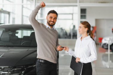 Gülümseyen adam sevinçle yumruğunu kaldırıyor, modern otomobil galerisindeki bir bayandan araba anahtarı alıyor.