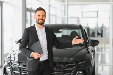 Profesyonel araba satıcısı gülümsüyor, pano tutuyor ve bir otomobil galerisinde yeni siyah bir arabaya doğru hareket ediyor.