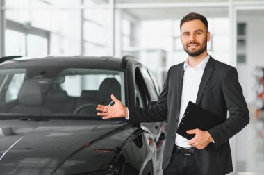 Galeride bir otomobilin yanında duran genç profesyonel araba satıcısı elinde bir pano ve gülümsüyor.