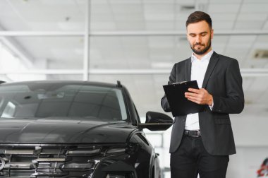 Satış elemanı elinde bir pano, parlak bir otomobil galerisindeki yeni siyah bir arabanın yanındaki belgeleri inceliyor.