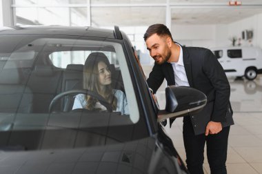 Yeni bir otomobilin içinde kadına yardım eden erkek, modern bir araba galerisinde araç satın alma sırasında özelliklerini tartışıyor.
