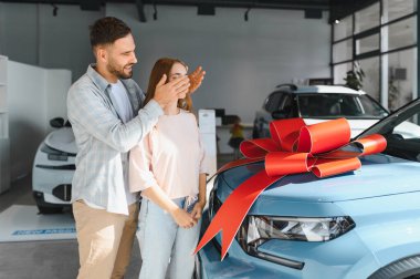 Yeni otomobil alımıyla sürpriz yapan bir adam. Gözleri ellerinde, büyük kırmızı kurdeleli mavi bir arabanın yanında duruyor.