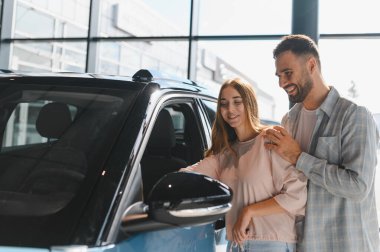 Gülümseyen genç bir çift sergilenen bir araca bakıyor, bir otomobil galerisinde satın alma kararı veriyorlar.