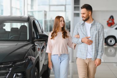 Genç bir çift bir otomobil galerisinde yürüyor, yeni arabalara bakıyor ve bir sonraki alışverişlerini düşünüyorlar.