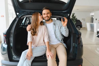 Gülümseyen genç bir çift yeni araba alımını kutluyor. Elinde anahtarlar olan bir adam. Bir kadın onu arabanın bagajında öpüyor.