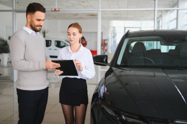 Araba seçeneklerini ve detayları modern bir otomobil galerisindeki erkek müşteriye gösteren satıcı, alışverişi tartışıyor.