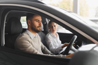 Erkek müşteri yeni bir arabanın içini incelerken, kadın satış elemanı ona bir galeride özelliklerini anlatıyor.