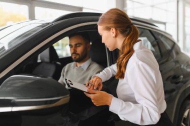 Satıcı kadın, bir otomobil galerisinde yeni bir arabanın içinde oturan bir erkek müşteriye sözleşmenin detaylarını sunuyor.