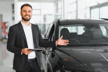 Otomobil galerisinde elinde bir dosya olan ve müşterilere siyah bir arabanın özelliklerini gösteren ve açıklayan bir erkek uzman.