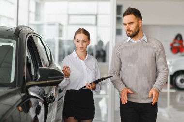 Araba galerisinde şüpheli bir erkek müşteriye yeni bir araç sunan genç satıcı, özellikleri ve fiyatları tartışıyor.