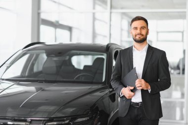 Profesyonel bir satıcı elinde bir dosya, modern bir galeride siyah bir arabanın yanında duruyor, araba satıyor.