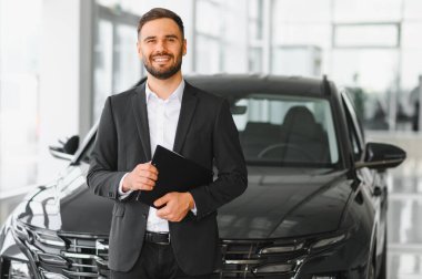 Elinde bir dosya olan takım elbiseli gülümseyen bir araba satıcısı, yeni bir araçla modern bir araba galerisinde duruyor.