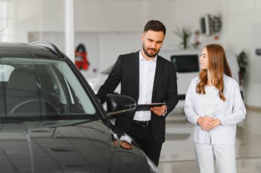 Modern bir galeride kadın müşteriye yeni araba özellikleri sunan satıcı seçenekleri tartışıyor ve ayrıntıları satın alıyor.