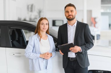 Satış elemanı ve bayan müşteri yeni arabanın yanında duruyor, satın alma seçeneklerini tartışıyor ve danışma sırasında gülümsüyorlar.