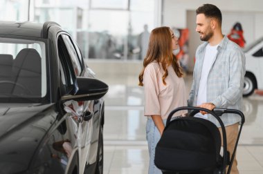 Bebek arabalı genç bir çift gülümsüyor ve modern bir galeride yeni bir araba seçiyorlar. Ailelerinin gelecekteki alışverişini planlıyorlar.