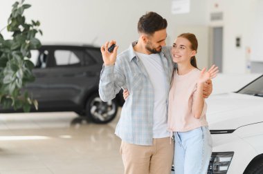 Hevesli genç çift araba galerisinde başarılı araba alışverişini kutluyor ve ellerinde yeni araba anahtarları tutuyorlar.