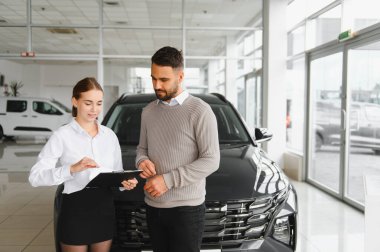 Profesyonel satış elemanı bir erkek müşteriyle evrak ve araba özelliklerini tartışıyor. Modern bir galeride bir araç satın alma anlaşmasını sonuçlandırıyor.