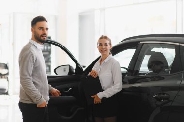 Bir araba galerisinde kadın ve erkek satış elemanı, araç seçenekleri ve alımları tartışıyor, müşteri hizmetlerini ve otomotiv perakendecilerini temsil ediyor.