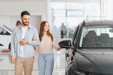 Araba galerisinde yeni bir araca bakan genç bir çift. Kadın ve erkek araba almayı düşünüyor, gülümsüyor.