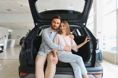 Genç çift kucaklaşıyorlar, yeni bir SUV 'un açık bagajında oturuyorlar. Parlak bir araba galerisinde. Araç alımlarını kutluyorlar.