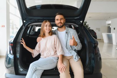 Yeni bir araç almalarını kutlayan mutlu genç bir çift, bir araba galerisindeki siyah ciplerinin bagajında oturuyorlar.