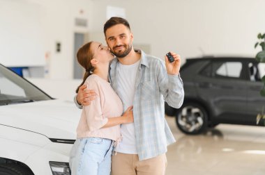 Gülümseyen genç çift kucaklaşıyorlar, yeni bir araba almalarını kutluyorlar, galerinin anahtarlarını tutuyorlar.